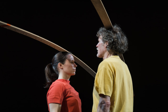 A woman with brown hair and a red shirt and a man with salt and pepper hair with a yellow shirt stand looking at each other. Each perches a thin wooden arc atop their heads.